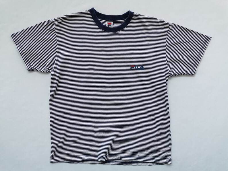 Puede incluir: Camiseta a rayas blancas y azul marino con cuello azul marino y el logo de Fila bordado en el pecho. La camiseta de manga corta tiene un dise&ntilde;o cl&aacute;sico e informal.