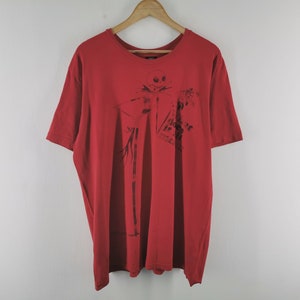 May include: Red t-shirt featuring a black graphic of Jack Skellington from The Nightmare Before Christmas. The design includes the text "Does it scare ya?". The shirt is hanging on a wooden hanger.