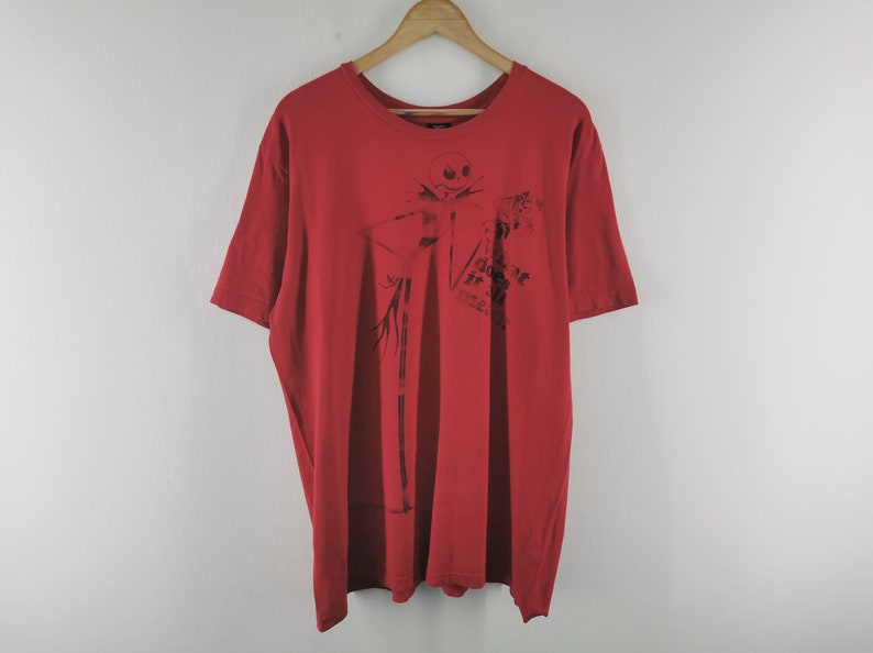 May include: Red t-shirt featuring a black graphic of Jack Skellington from The Nightmare Before Christmas. The design includes the text "Does it scare ya?". The shirt is hanging on a wooden hanger.