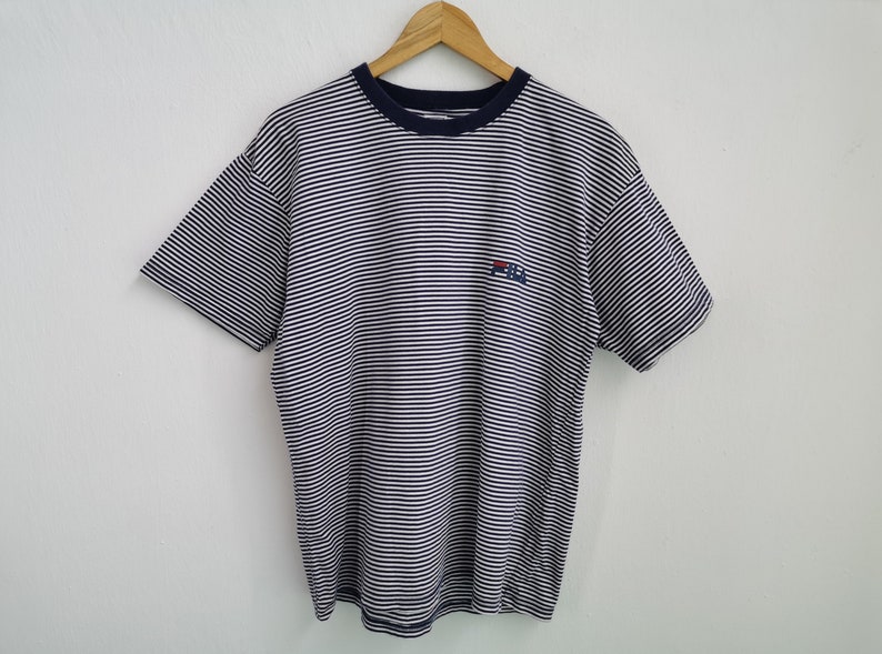 Puede incluir: Camiseta a rayas blancas y azul marino con cuello y mangas cortas azul marino. Tiene un peque&ntilde;o logotipo en el pecho. La camiseta est&aacute; colgada de una percha de madera sobre un fondo blanco.