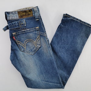 May include: Blue denim jeans with a faded wash. The jeans feature a unique pocket design with decorative stitching and a small red tag. The waistband has a brown leather patch with the text "Blue Trip".