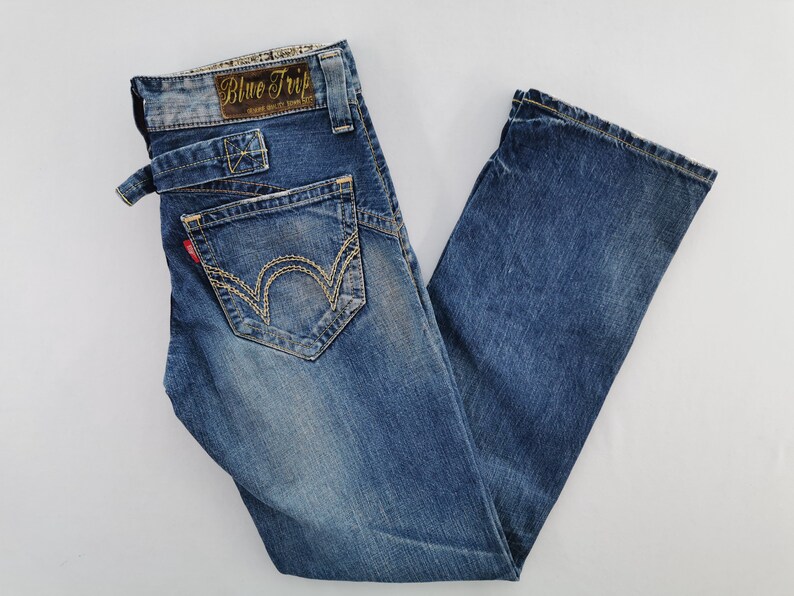 May include: Blue denim jeans with a faded wash. The jeans feature a unique pocket design with decorative stitching and a small red tag. The waistband has a brown leather patch with the text "Blue Trip".