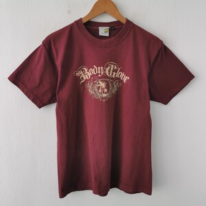 May include: A maroon t-shirt featuring the text "Body Glove" in a decorative font, with a palm tree and shield graphic. The shirt is displayed on a wooden hanger against a white wall.