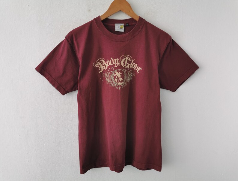May include: A maroon t-shirt featuring the text "Body Glove" in a decorative font, with a palm tree and shield graphic. The shirt is displayed on a wooden hanger against a white wall.