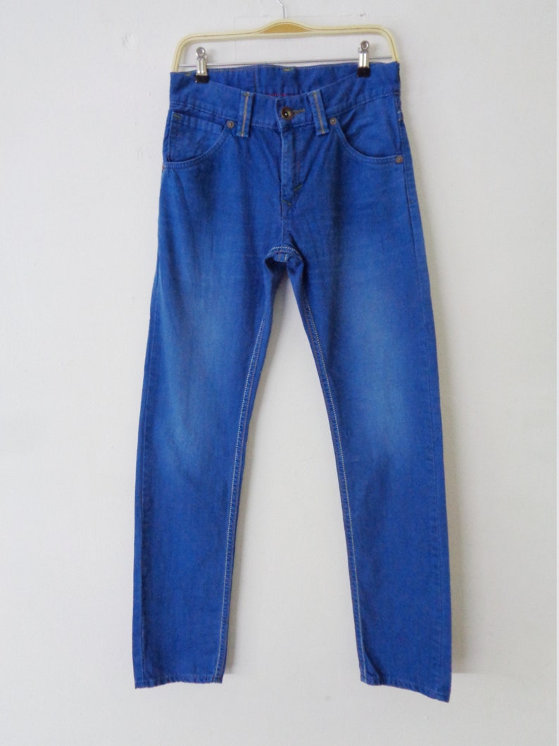 K&ouml;nnte beinhalten: Hellblaue Jeans aus Denim, die an einem Kleiderb&uuml;gel h&auml;ngen. Die Jeans haben ein klassisches F&uuml;nf-Taschen-Design mit Knopf- und Rei&szlig;verschluss. Die N&auml;hte sind wei&szlig;, und der Denim hat ein leicht verwaschenes Aussehen.