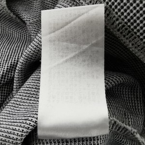 May include: A white rectangular garment care label with printed text attached to a black and white micro-check patterned fabric. The label is vertically oriented and features multiple lines of small, light-colored text printed on the surface.