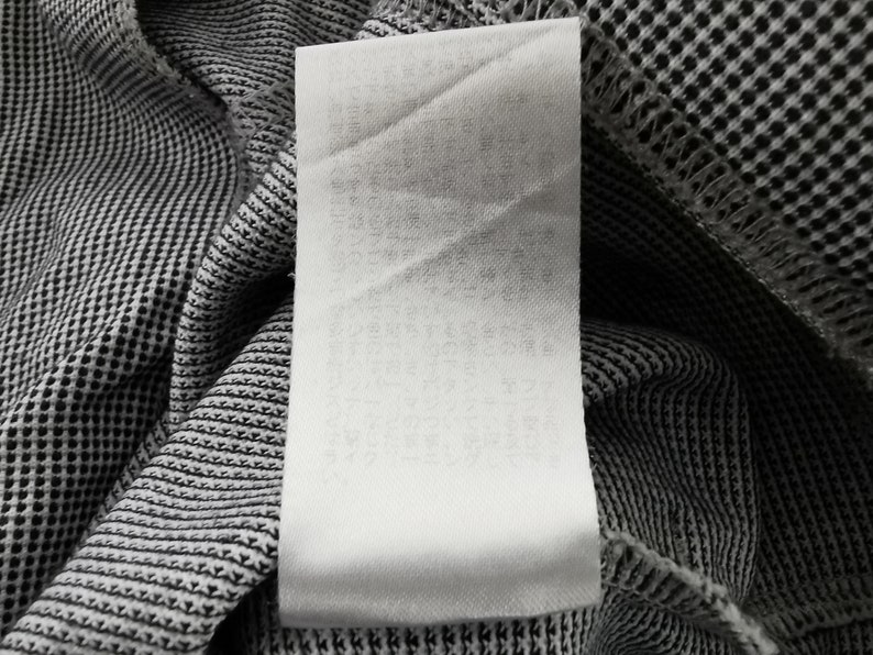 May include: A white rectangular garment care label with printed text attached to a black and white micro-check patterned fabric. The label is vertically oriented and features multiple lines of small, light-colored text printed on the surface.