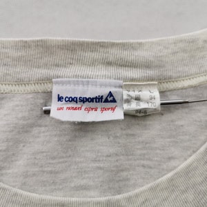 May include: Close-up of a light grey t-shirt collar with a sewn-in label. The label reads "le coq sportif" with a rooster logo and French text. Japanese text and measurements are also visible.
