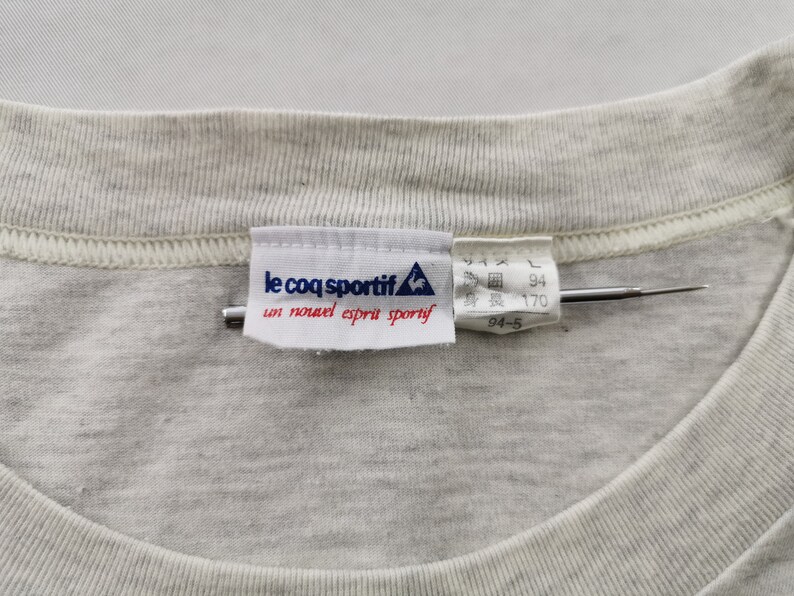 May include: Close-up of a light grey t-shirt collar with a sewn-in label. The label reads "le coq sportif" with a rooster logo and French text. Japanese text and measurements are also visible.