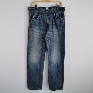 May include: Blue denim jeans with a faded wash and distressed details. The jeans are hanging on a wooden hanger against a white background. The jeans have a classic five-pocket design and a straight leg.