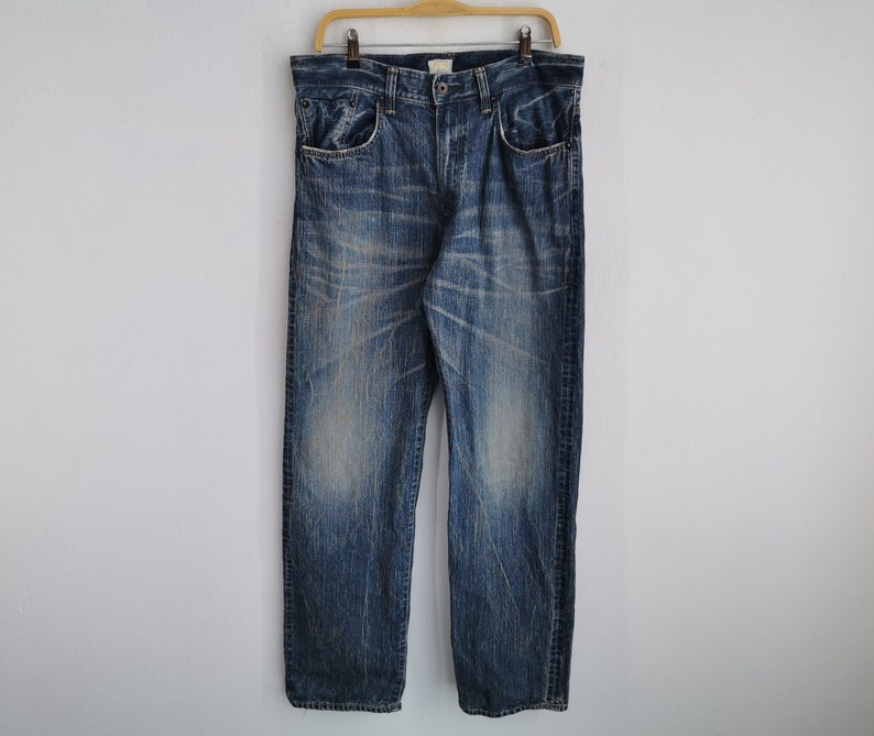 May include: Blue denim jeans with a faded wash and distressed details. The jeans are hanging on a wooden hanger against a white background. The jeans have a classic five-pocket design and a straight leg.