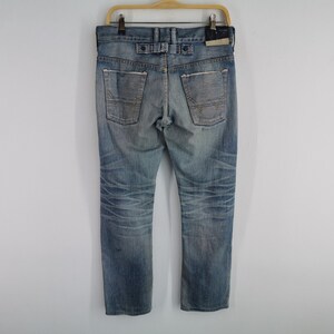 K&ouml;nnte beinhalten: Verblasste blaue Jeans mit Used-Look, mit Knopfverschluss und Details an den Ges&auml;&szlig;taschen. Die Jeans h&auml;ngen an einem Holzb&uuml;gel vor wei&szlig;em Hintergrund. Der Denim hat ein abgenutztes Aussehen.