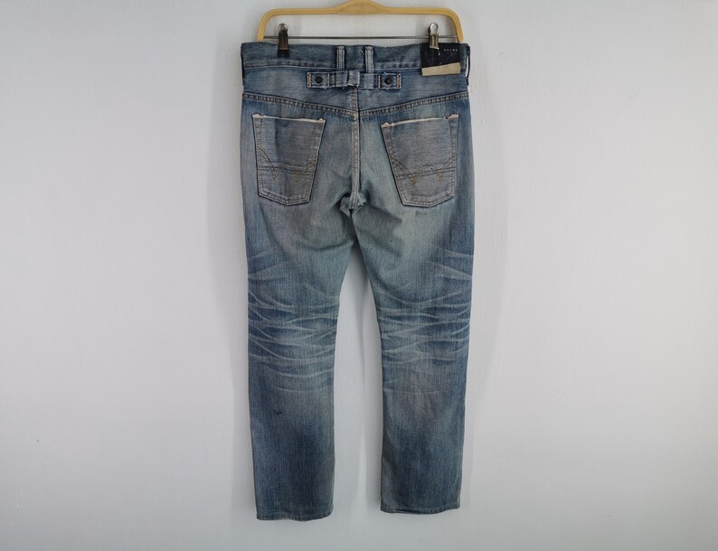 K&ouml;nnte beinhalten: Verblasste blaue Jeans mit Used-Look, mit Knopfverschluss und Details an den Ges&auml;&szlig;taschen. Die Jeans h&auml;ngen an einem Holzb&uuml;gel vor wei&szlig;em Hintergrund. Der Denim hat ein abgenutztes Aussehen.