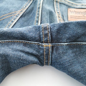 May include: Close-up of blue denim jeans with visible stitching in a contrasting tan color. The Wrangler brand label is visible. The jeans have a classic five-pocket design.