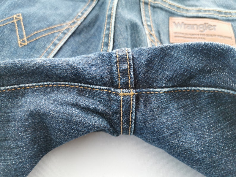 May include: Close-up of blue denim jeans with visible stitching in a contrasting tan color. The Wrangler brand label is visible. The jeans have a classic five-pocket design.