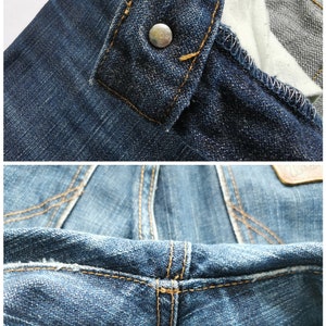 May include: Detailed view of blue denim jeans, highlighting the button fastening and stitching. The jeans feature a traditional design with prominent seams and a metal button. The fabric has a worn, textured appearance.