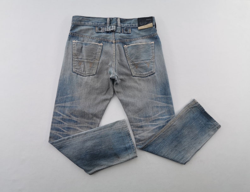K&ouml;nnte beinhalten: Hellblaue Jeans mit verwaschener Optik und Used-Details. Die Jeans haben ein klassisches F&uuml;nf-Taschen-Design, einen Knopf- und Rei&szlig;verschluss und einen Lederaufn&auml;her auf der R&uuml;ckseite. Die Jeans liegen flach auf einer wei&szlig;en Oberfl&auml;che.