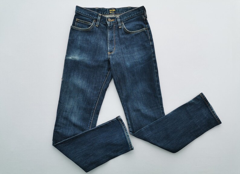 May include: Dark blue denim jeans with a classic five-pocket design. The jeans have a button and zipper closure, belt loops, and a faded wash. The Wrangler logo is visible on the back pocket. A timeless wardrobe staple.