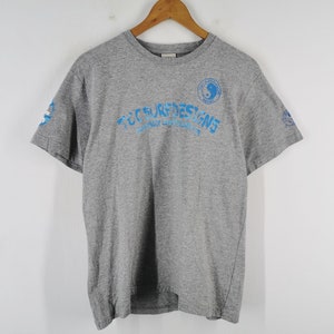 May include: A gray short-sleeve t-shirt with blue text and graphics. The text reads "TCC SURF DESIGNS" in a curved font. A yin-yang symbol is on the right chest. Additional graphics are on the sleeves. The shirt is hanging on a wooden hanger.