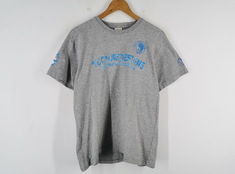 May include: A gray short-sleeve t-shirt with blue text and graphics. The text reads "TCC SURF DESIGNS" in a curved font. A yin-yang symbol is on the right chest. Additional graphics are on the sleeves. The shirt is hanging on a wooden hanger.