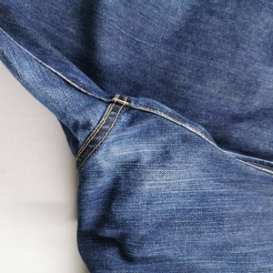 May include: Close-up of blue denim jeans. The jeans feature yellow stitching along the seams and pockets. The fabric has a textured appearance, with visible wear and fading. The image highlights the details of the garment's construction.