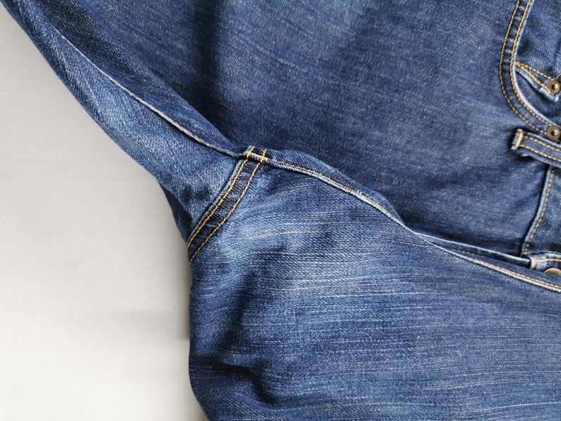 May include: Close-up of blue denim jeans. The jeans feature yellow stitching along the seams and pockets. The fabric has a textured appearance, with visible wear and fading. The image highlights the details of the garment's construction.