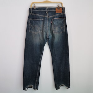 Pu&ograve; includere: Jeans di denim blu con un lavaggio sbiadito, appesi a una gruccia di legno. I jeans hanno un classico design a cinque tasche, una toppa in pelle e dettagli strappati sull'orlo. Le tasche posteriori presentano un motivo di cucitura distintivo.
