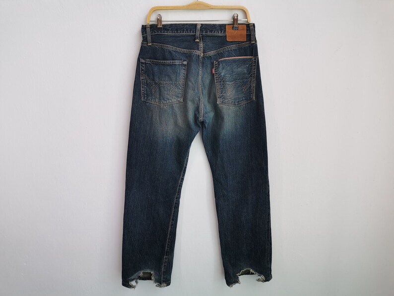 Pu&ograve; includere: Jeans di denim blu con un lavaggio sbiadito, appesi a una gruccia di legno. I jeans hanno un classico design a cinque tasche, una toppa in pelle e dettagli strappati sull'orlo. Le tasche posteriori presentano un motivo di cucitura distintivo.