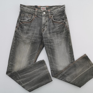 May include: Faded black denim jeans with a button closure and classic five-pocket design. The jeans have orange stitching details and a slightly distressed appearance, suggesting a vintage or worn aesthetic. The jeans are laid flat on a white surface.