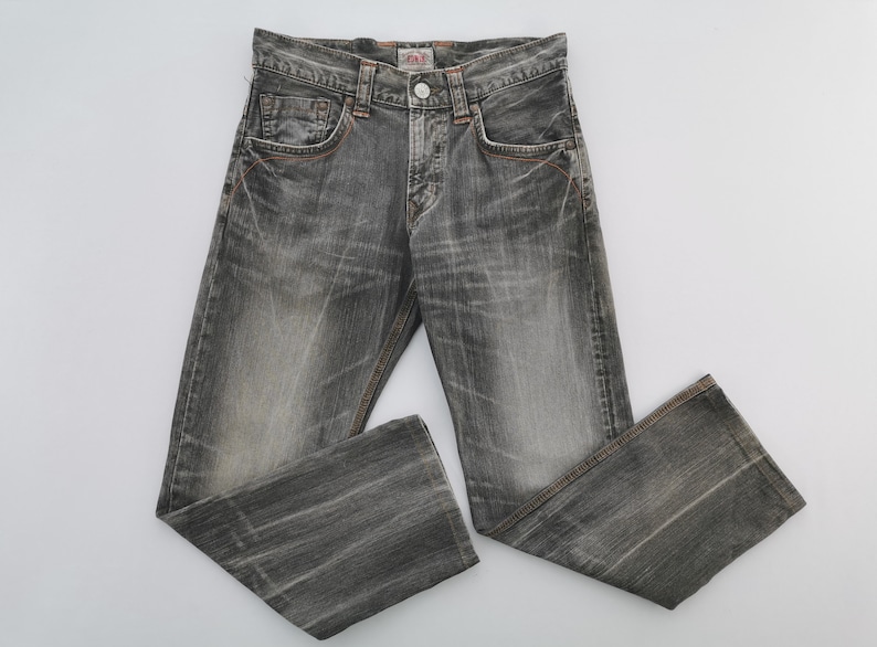 May include: Faded black denim jeans with a button closure and classic five-pocket design. The jeans have orange stitching details and a slightly distressed appearance, suggesting a vintage or worn aesthetic. The jeans are laid flat on a white surface.