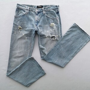 Pu&ograve; includere: Jeans in denim blu chiaro con dettagli strappati, tra cui sezioni strappate sul davanti. I jeans hanno un classico design a cinque tasche e una vestibilit&agrave; dritta. I jeans sono stesi su una superficie bianca.