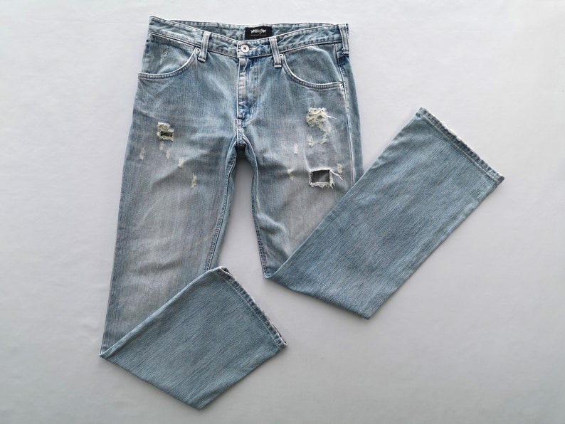 Pu&ograve; includere: Jeans in denim blu chiaro con dettagli strappati, tra cui sezioni strappate sul davanti. I jeans hanno un classico design a cinque tasche e una vestibilit&agrave; dritta. I jeans sono stesi su una superficie bianca.