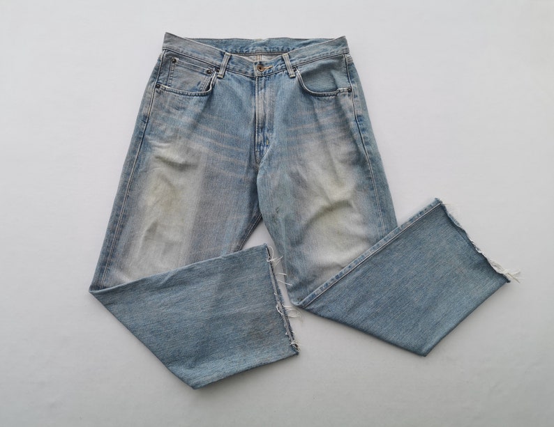 Puede incluir: Vaqueros de mezclilla azul claro con un lavado descolorido y detalles desgastados. Los vaqueros tienen un dise&ntilde;o cl&aacute;sico de cinco bolsillos, cierre de bot&oacute;n y cremallera, y una pernera recta con un dobladillo sin rematar y deshilachado. Los vaqueros est&aacute;n planos sobre una superficie blanca.
