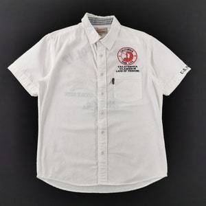 May include: White short-sleeved shirt with a button-down collar and a chest pocket. Features a red and white circular graphic with the text "CALIFORNIA SURF LIFE" and "CALIFORNIA CLASSICS LAND OF PROMISE". The sleeve has "U.S.N" printed on it.