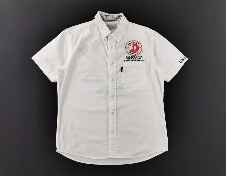 May include: White short-sleeved shirt with a button-down collar and a chest pocket. Features a red and white circular graphic with the text "CALIFORNIA SURF LIFE" and "CALIFORNIA CLASSICS LAND OF PROMISE". The sleeve has "U.S.N" printed on it.