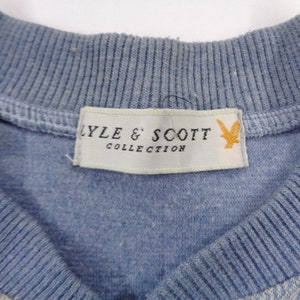 Puede incluir: Primer plano de un su&eacute;ter azul claro con cuello de canal&eacute;. La etiqueta dice "LYLE & SCOTT COLLECTION" con un logotipo de p&aacute;jaro amarillo. El su&eacute;ter tiene un patr&oacute;n sutil en la parte inferior.