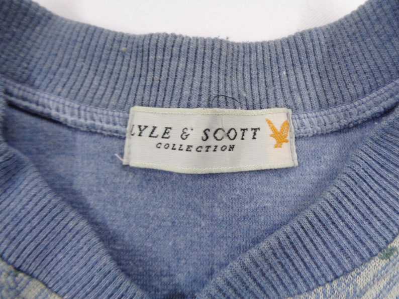 Puede incluir: Primer plano de un su&eacute;ter azul claro con cuello de canal&eacute;. La etiqueta dice "LYLE & SCOTT COLLECTION" con un logotipo de p&aacute;jaro amarillo. El su&eacute;ter tiene un patr&oacute;n sutil en la parte inferior.