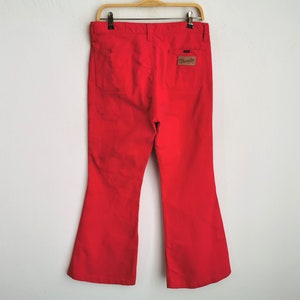 May include: Bright red Wrangler bell-bottom pants hanging on a wooden hanger. The pants have two back pockets and a brown leather patch with the Wrangler logo. The pants are made of a sturdy material and have a flared leg opening.