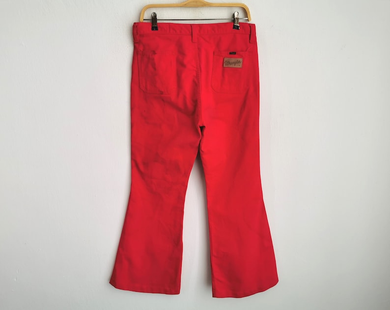 May include: Bright red Wrangler bell-bottom pants hanging on a wooden hanger. The pants have two back pockets and a brown leather patch with the Wrangler logo. The pants are made of a sturdy material and have a flared leg opening.