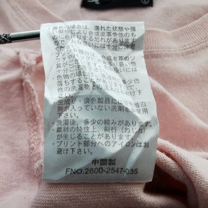 May include: Close-up of a light pink t-shirt with a white tag attached. The tag contains Japanese text and the number "2600-2547-035". The shirt's fabric is visible, showing the texture of the material.