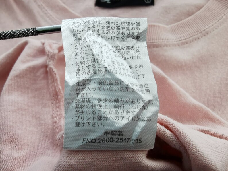 May include: Close-up of a light pink t-shirt with a white tag attached. The tag contains Japanese text and the number "2600-2547-035". The shirt's fabric is visible, showing the texture of the material.