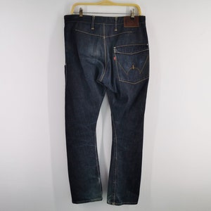 May include: Dark wash denim jeans with a classic five-pocket design. The jeans have a brown leather patch on the back and a red tag on the back pocket. The jeans are hanging on a wooden hanger against a white background.
