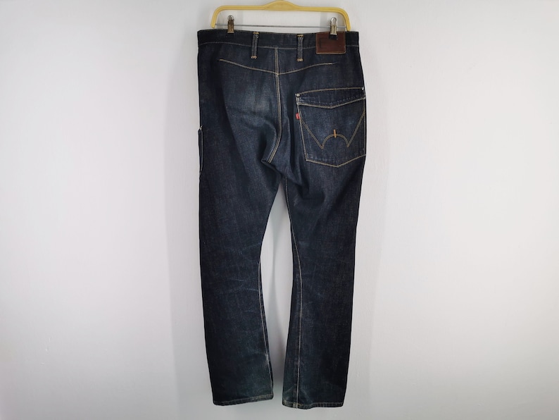 May include: Dark wash denim jeans with a classic five-pocket design. The jeans have a brown leather patch on the back and a red tag on the back pocket. The jeans are hanging on a wooden hanger against a white background.