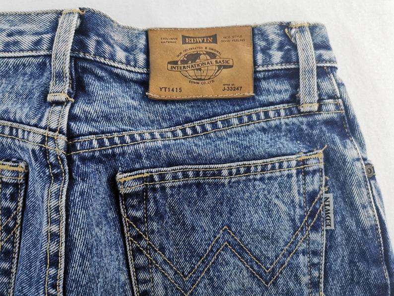 Puede incluir: Primer plano de unos vaqueros de mezclilla azules con un lavado descolorido. Los bolsillos traseros presentan un dise&ntilde;o distintivo y la etiqueta de la marca "EDWIN INTERNATIONAL BASIC" es visible. Los vaqueros tienen una est&eacute;tica cl&aacute;sica y vintage.