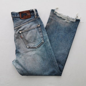 Puede incluir: Vaqueros de mezclilla azul claro con un dobladillo deshilachado y deshilachado. Los jeans presentan un dise&ntilde;o cl&aacute;sico de cinco bolsillos, un parche de cuero marr&oacute;n y una etiqueta roja. Los jeans est&aacute;n doblados, mostrando los bolsillos traseros y la etiqueta de la marca.