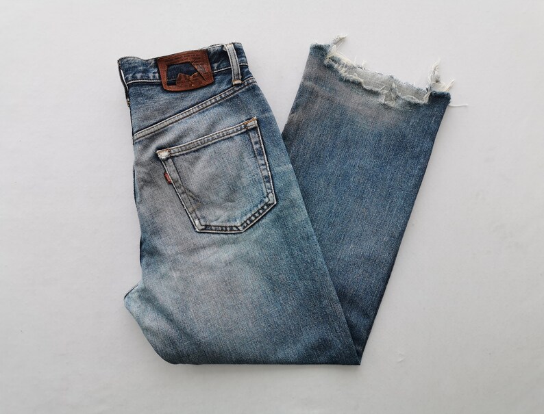Puede incluir: Vaqueros de mezclilla azul claro con un dobladillo deshilachado y deshilachado. Los jeans presentan un dise&ntilde;o cl&aacute;sico de cinco bolsillos, un parche de cuero marr&oacute;n y una etiqueta roja. Los jeans est&aacute;n doblados, mostrando los bolsillos traseros y la etiqueta de la marca.