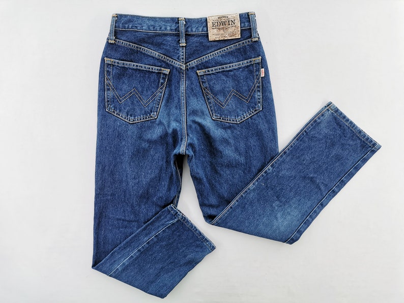 Puede incluir: Vaqueros de mezclilla azul oscuro con un dise&ntilde;o cl&aacute;sico de cinco bolsillos. Presentan cintura alta, pernera recta y el nombre de la marca "EDWIN" en un parche de cuero. Los bolsillos traseros tienen una costura distintiva en forma de W.