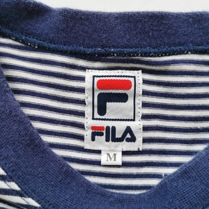 Puede incluir: Primer plano de una camiseta a rayas azul marino y blanca con cuello en V. La camiseta presenta una etiqueta de la marca Fila con un logotipo rojo y blanco y el texto "FILA". La etiqueta tambi&eacute;n indica la talla "M".