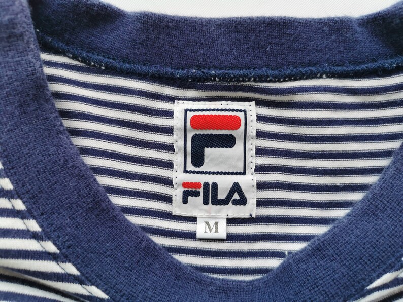 Puede incluir: Primer plano de una camiseta a rayas azul marino y blanca con cuello en V. La camiseta presenta una etiqueta de la marca Fila con un logotipo rojo y blanco y el texto "FILA". La etiqueta tambi&eacute;n indica la talla "M".
