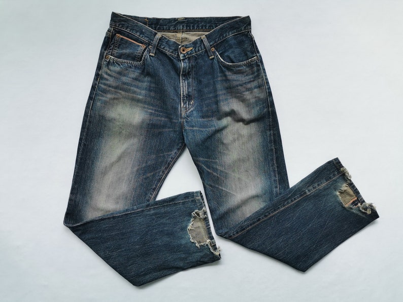 Pu&ograve; includere: Jeans in denim blu sbiadito con dettagli distressed. I jeans hanno un classico design a cinque tasche, una chiusura con bottone e cerniera e un aspetto usurato. Gli orli sono sfilacciati, aggiungendo all'estetica vintage. Un capo casual.